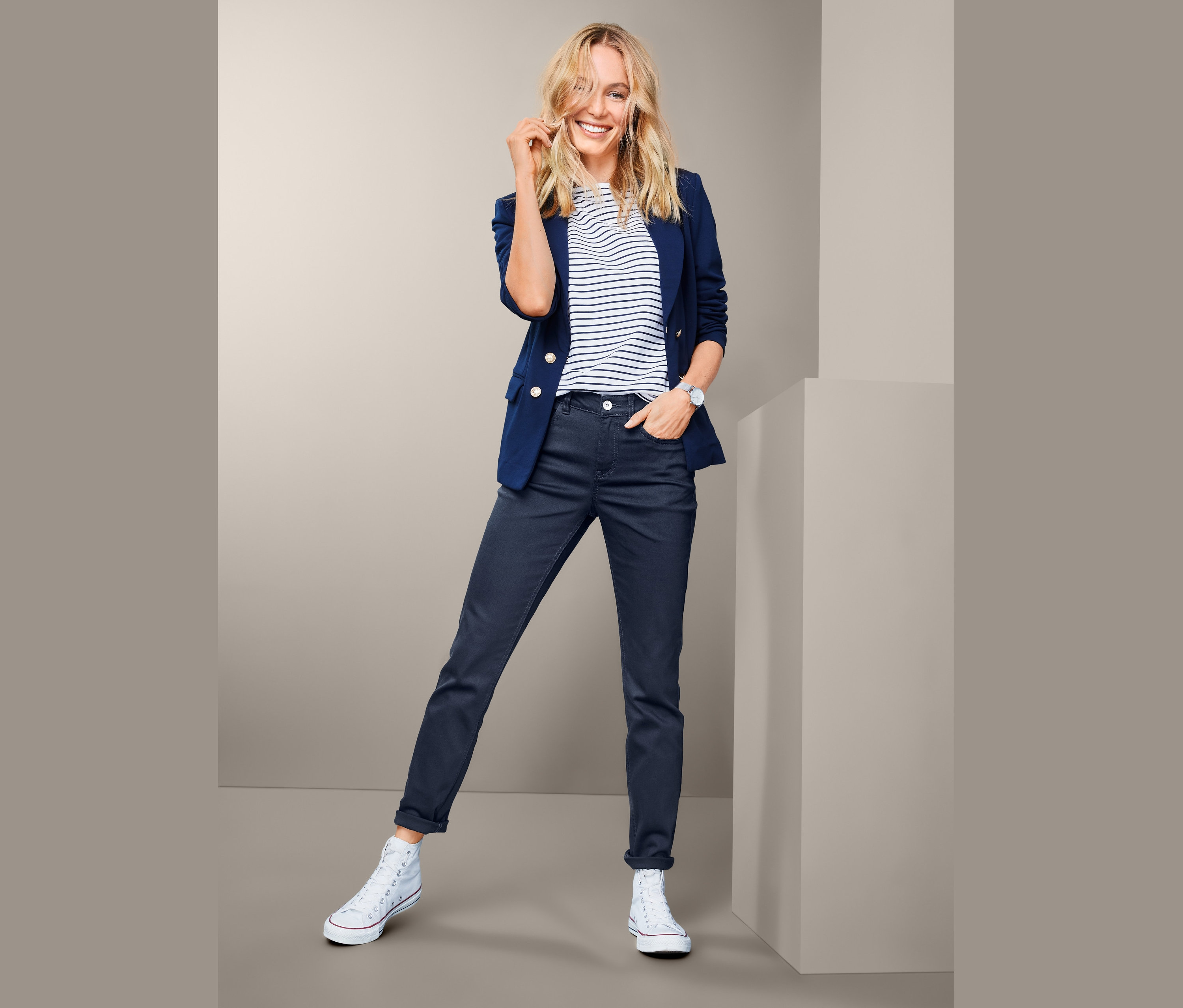 Blonde Frau posiert in marineblauem Blazer, gestreiftem T-Shirt, Jeans und weißen Schuhen.