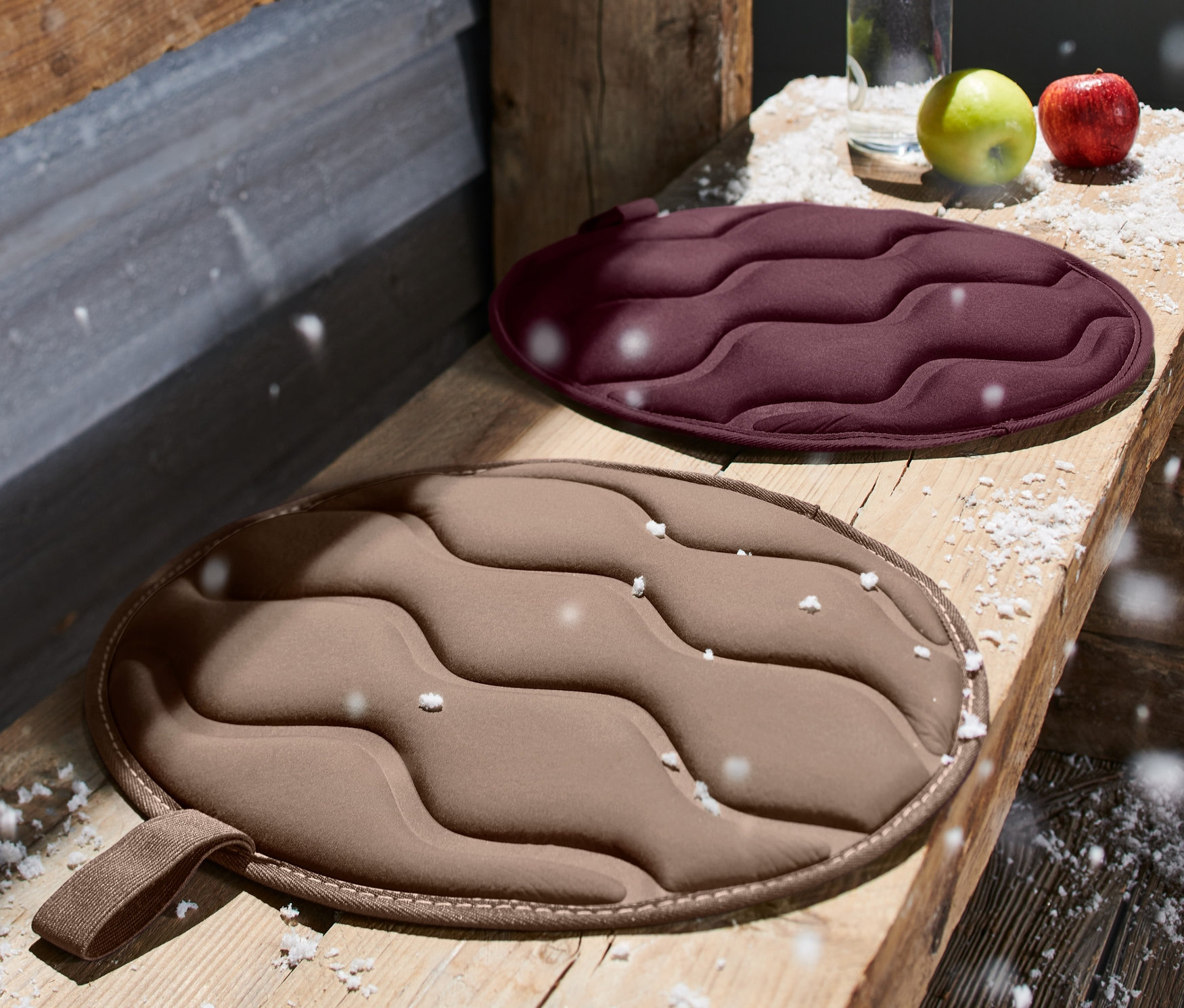 Zwei runde, faltbare Thermo-Sitzkissen liegen auf einer Holzbank. Eines ist bordeauxrot, das andere beige.