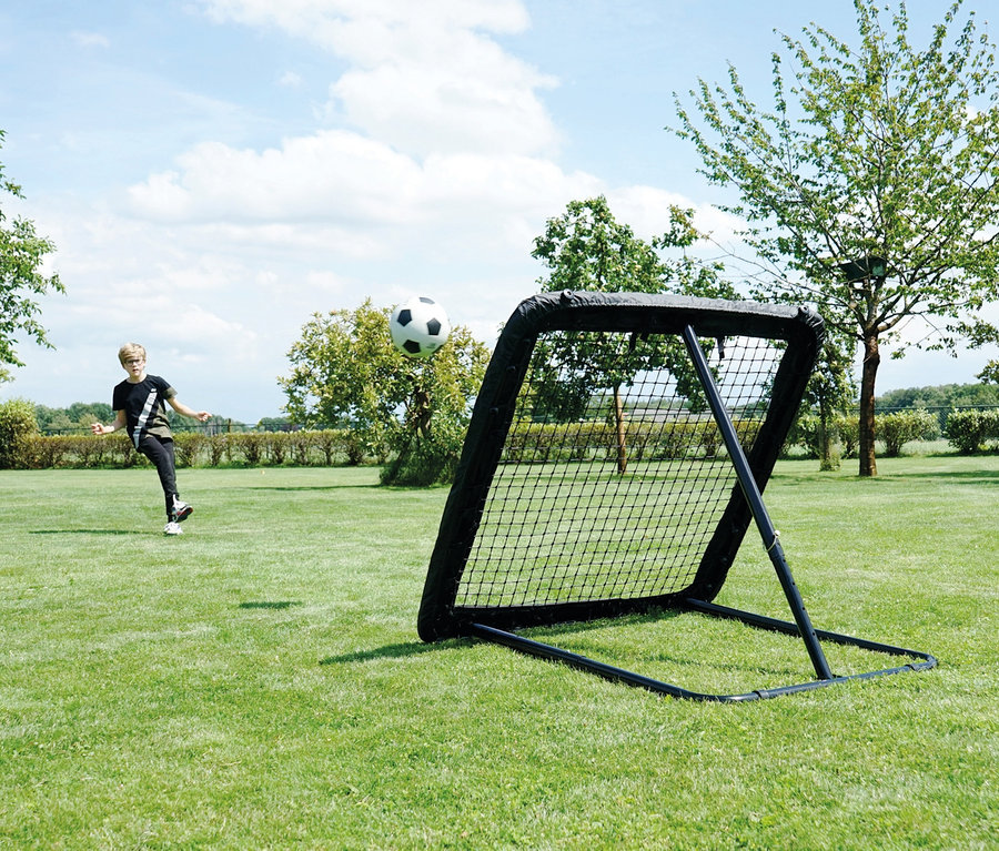 Ein Junge schießt einen Fußball in Richtung eines schwarzen EXIT Fußball-Rebounder L auf einer Wiese.