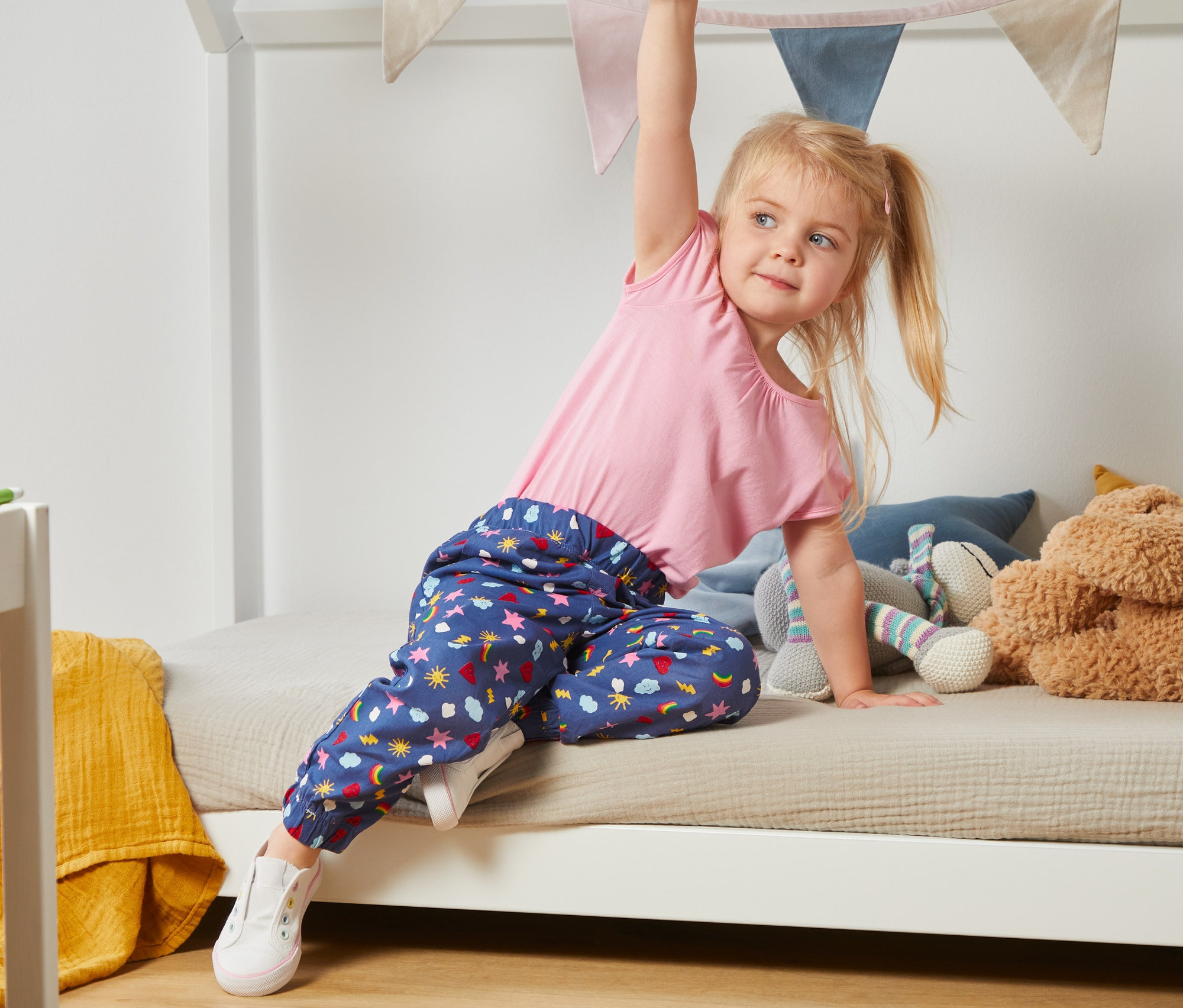 Mädchen in rosa Top und blau-bunter Schlupfhose aus Bio-Baumwolle klettert auf ein weißes Bett.