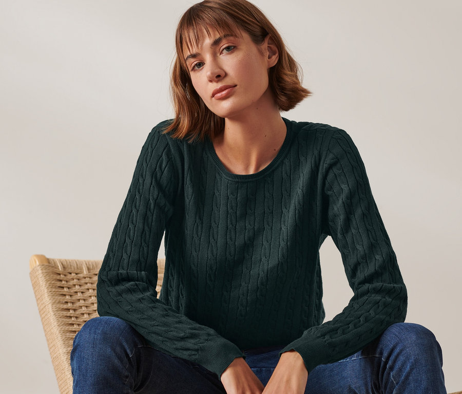 Frau sitzt auf einem Korbsessel und trägt einen dunkelgrünen Zopfstrickpullover.