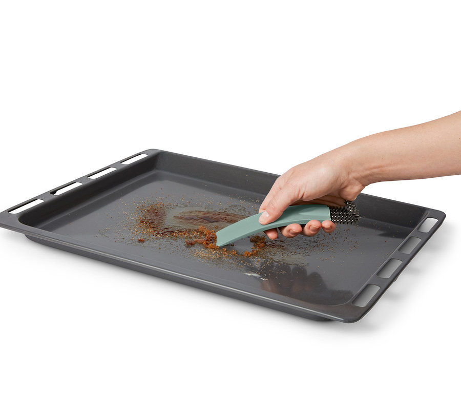 Eine Hand reinigt ein schmutziges Backblech mit einer Backofenbürste mit Schaber.
