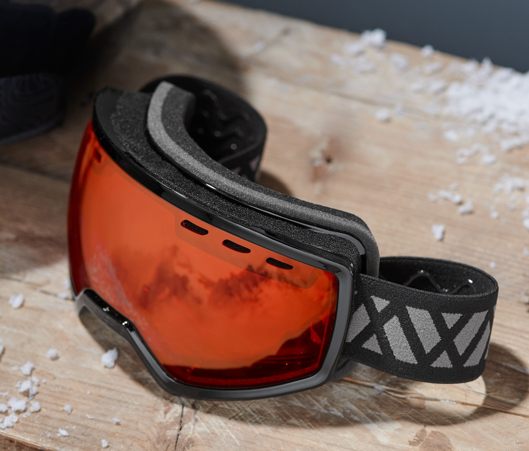 Eine Schneebrille mit orangefarbenem Glas und schwarzem Rahmen liegt auf einem Holztisch neben einem schwarzen Skihelm.