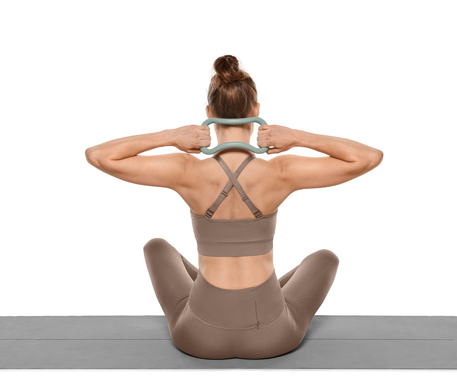 Frau sitzt auf Yogamatte und hält Yoga-Stretchring hinter ihrem Nacken.