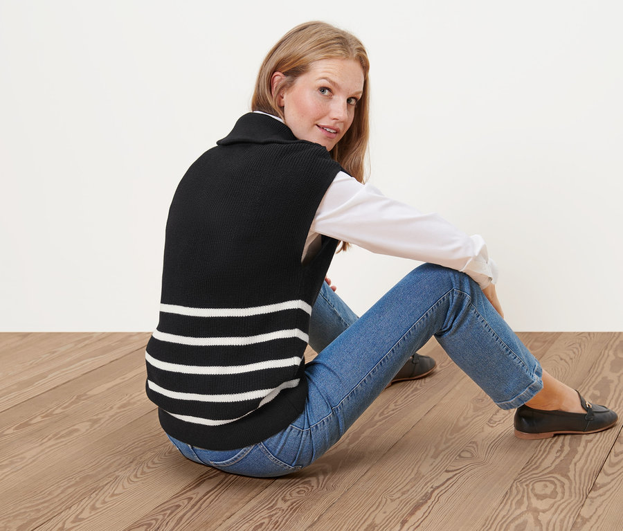 Frau sitzt auf dem Boden und trägt einen schwarzen Pullunder im Troyer-Stil mit weißen Streifen.