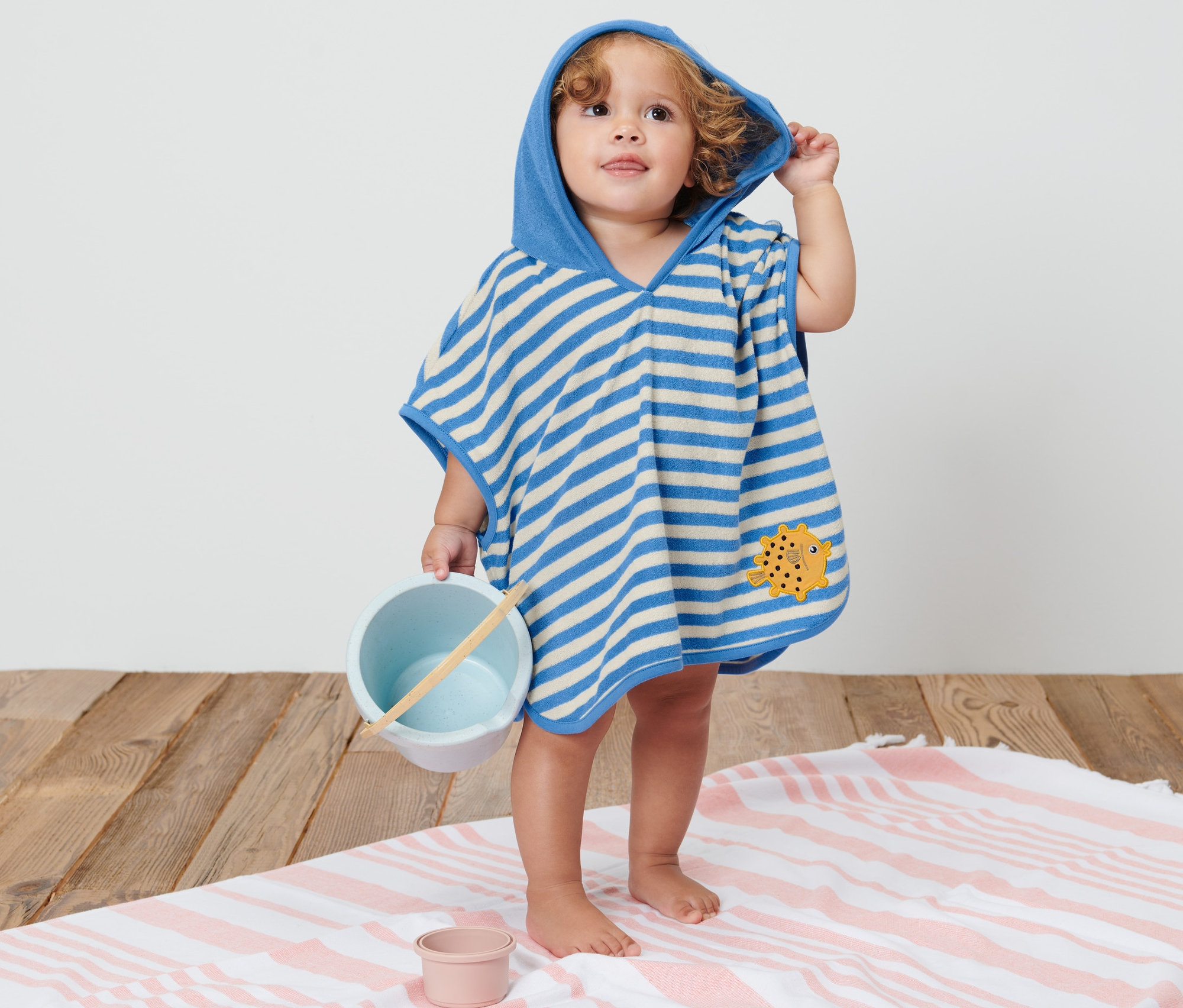 Ein Kind in einem blau-weiss gestreiften Kinder-Frottierponcho mit Kapuze hält ein Spielzeug.