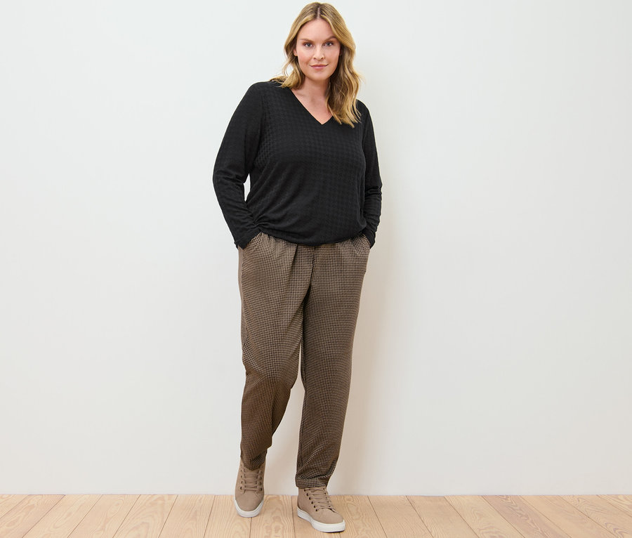 Eine Frau steht mit den Händen in den Taschen einer Joggpants mit Hahnentritt-Jacquard und einem schwarzen, strukturierten Langarmshirt.