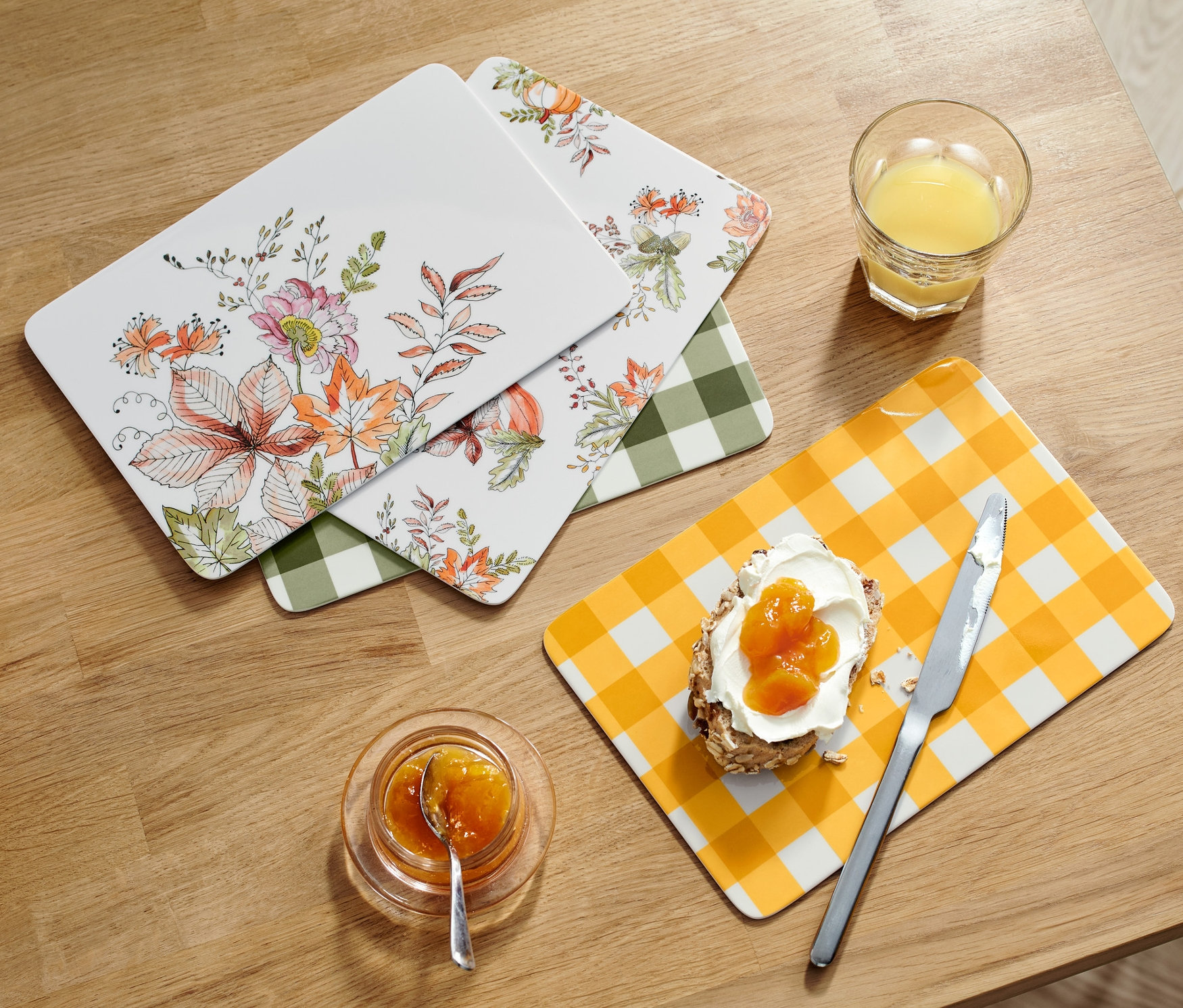Vier Frühstücksbretter liegen auf einem Holztisch neben einem Glas Saft, einem Brot mit Marmelade, Marmelade und einem Brotkorb.