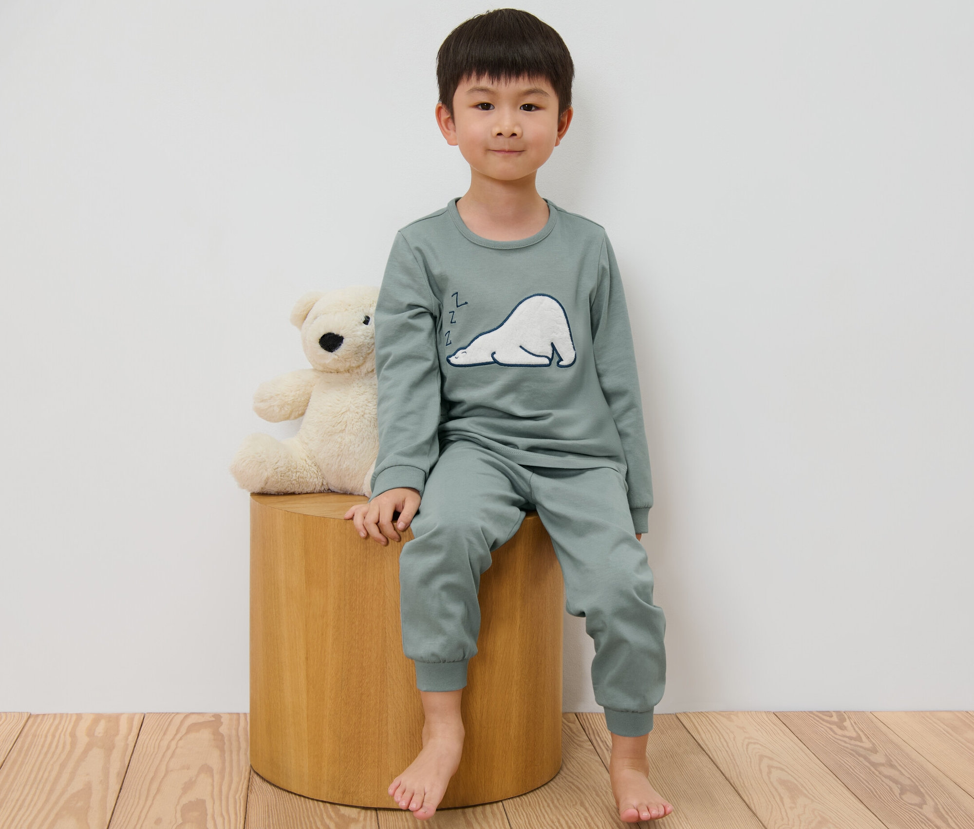 Junge sitzt auf Hocker neben Teddybär und trägt Kinder-Pyjama.