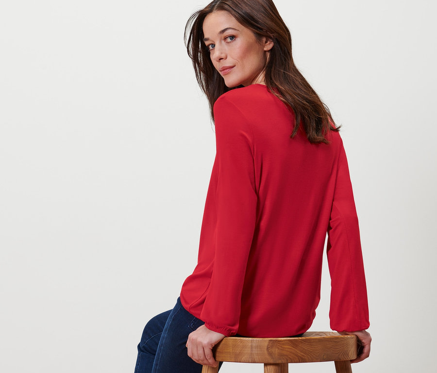 Frau sitzt auf einem Hocker und trägt ein rotes Blusenshirt.