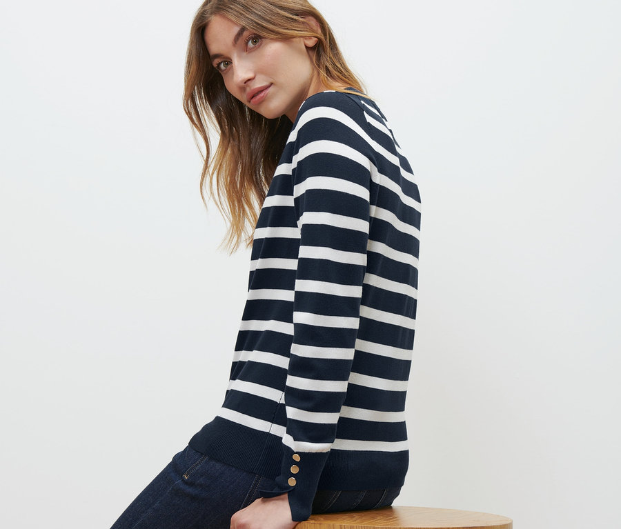 Frau sitzt auf einem Holzhocker und trägt einen gestreiften Feinstrickpullover in Navy und Jeans.