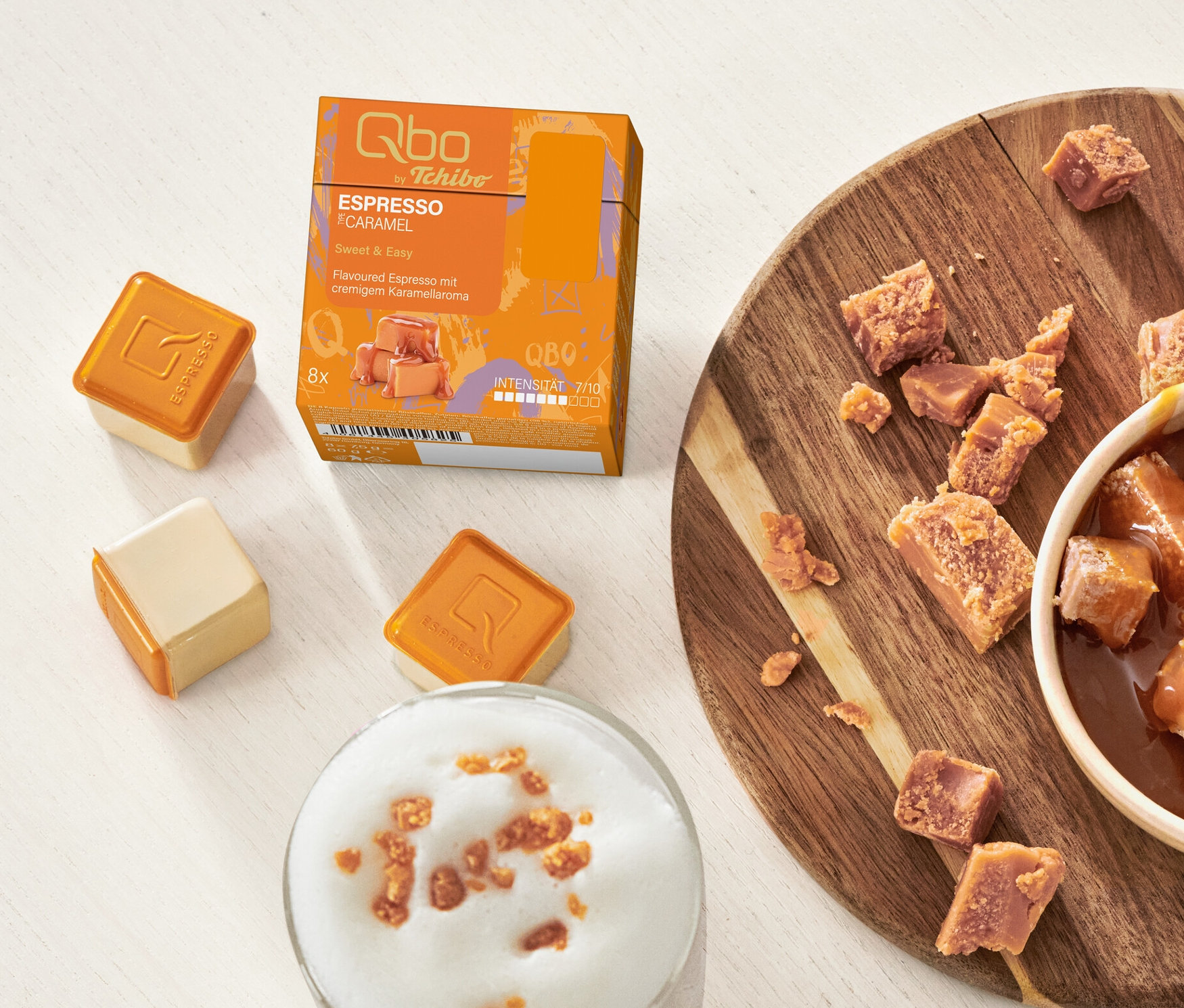 Auf einem Tisch stehen Qbo Flavoured Espresso Caramel Kapseln, eine Schale Karamellwürfel und ein Glas mit Karamell-Latte.