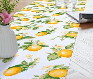 Dekorativer Tischläufer mit Zitronenmuster, Blumenvase, Wasserkaraffe und Glas auf Holztisch.