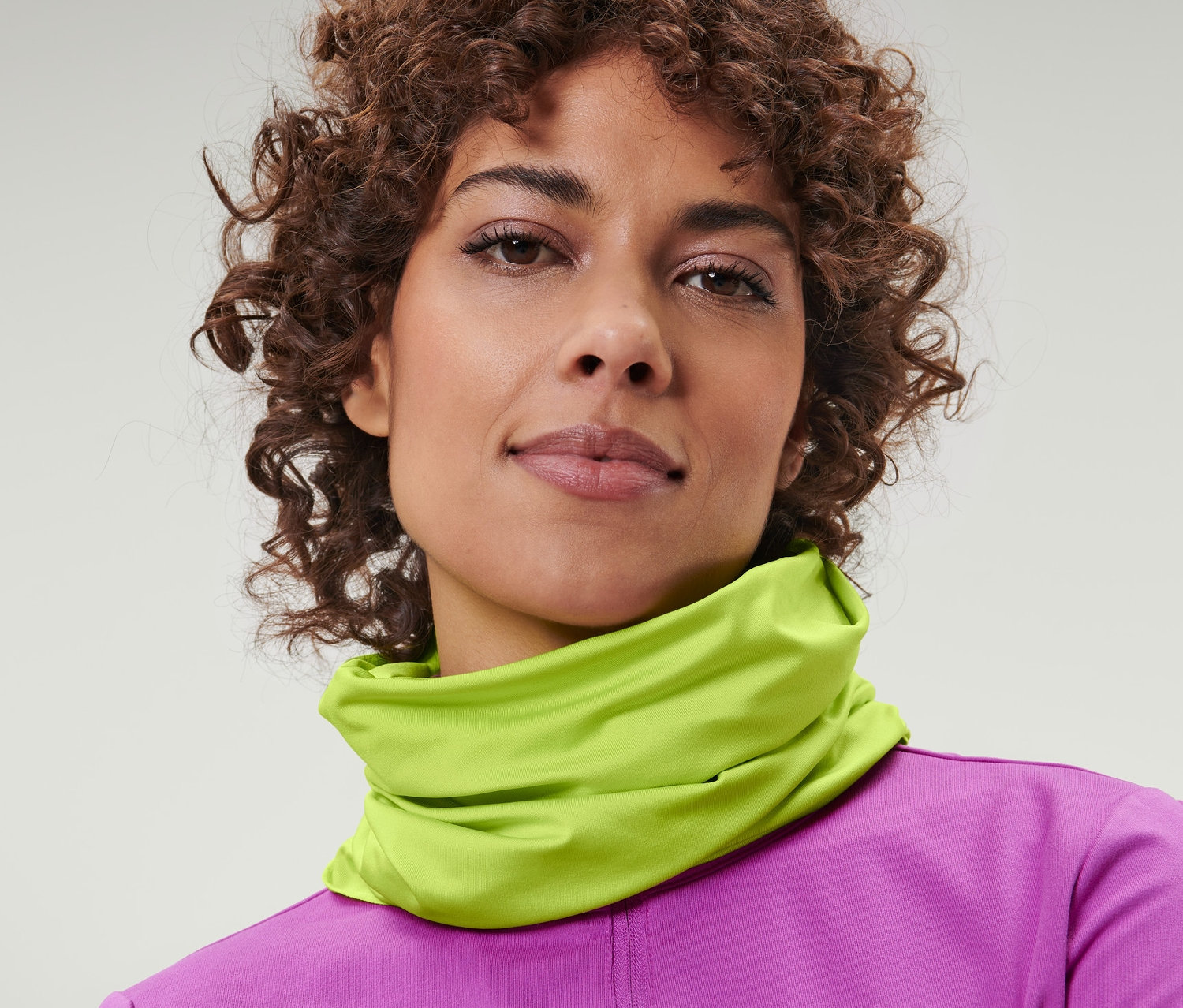 Porträt einer Frau mit braunen lockigen Haaren, die ein gelbes Multifunktionstuch um den Hals trägt.