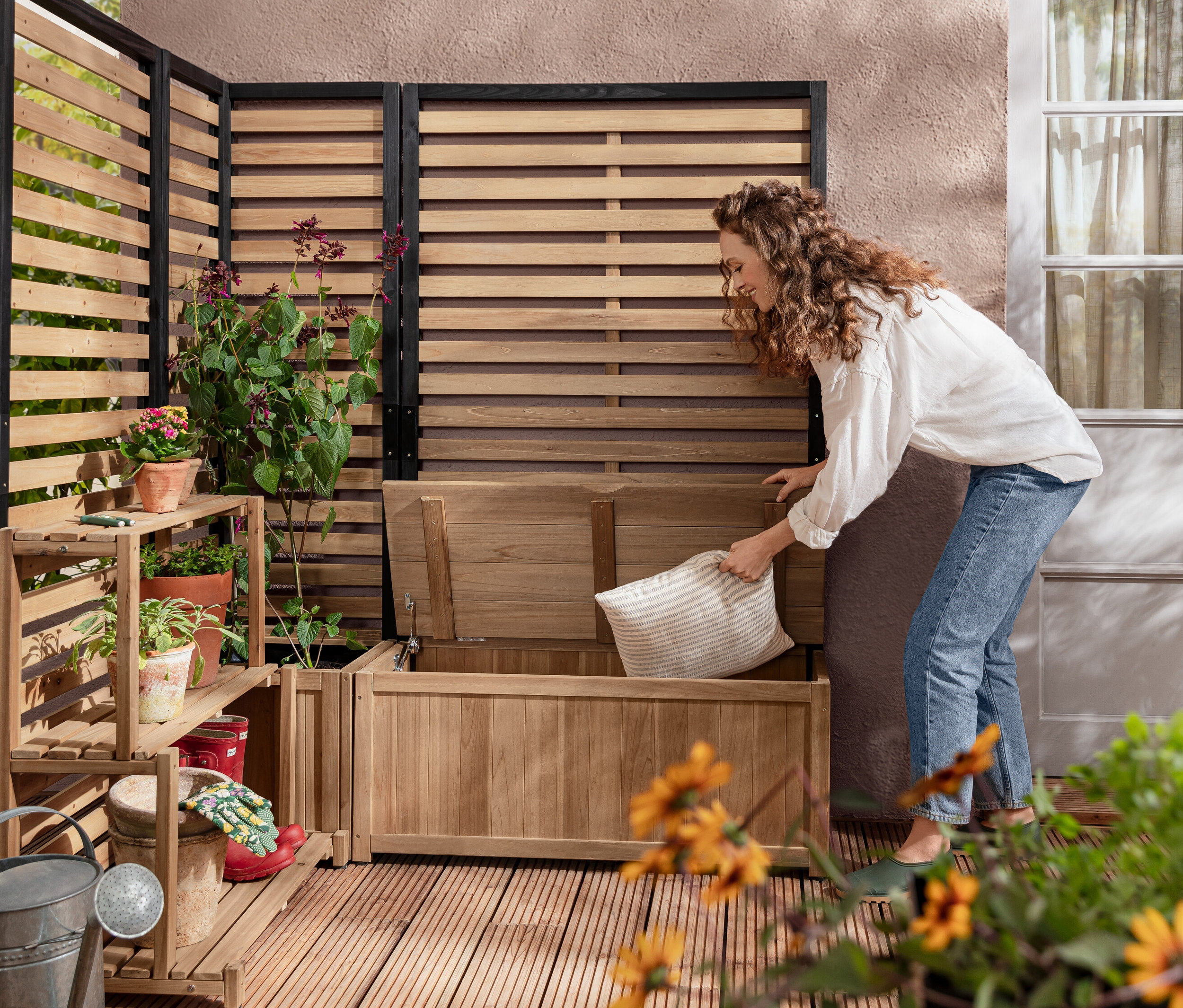 Eine Frau legt ein Kissen in die Sichtschutz-Stauraumbank-Modul »Elin«. Im Hintergrund befinden sich ein Sichtschutz-Pflanzkasten-Modul »Elin« und ein Sichtschutz-Regal-Modul »Elin«.