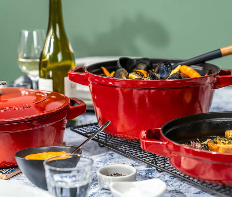 Auf einem Tisch stehen drei rote STAUB Kochtopf-Sets "La Cocotte". Ein Topf ist mit Muscheln gefüllt, ein anderer mit Croutons und der dritte mit Sauce.