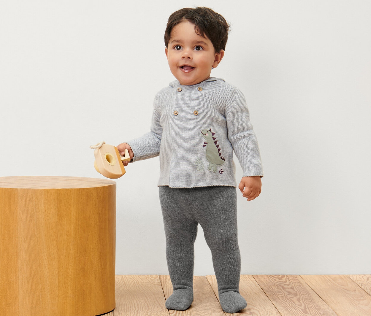 Ein Junge steht und hält ein Holzspielzeug, er trägt eine melierte Baby-Thermostrumpfhose.