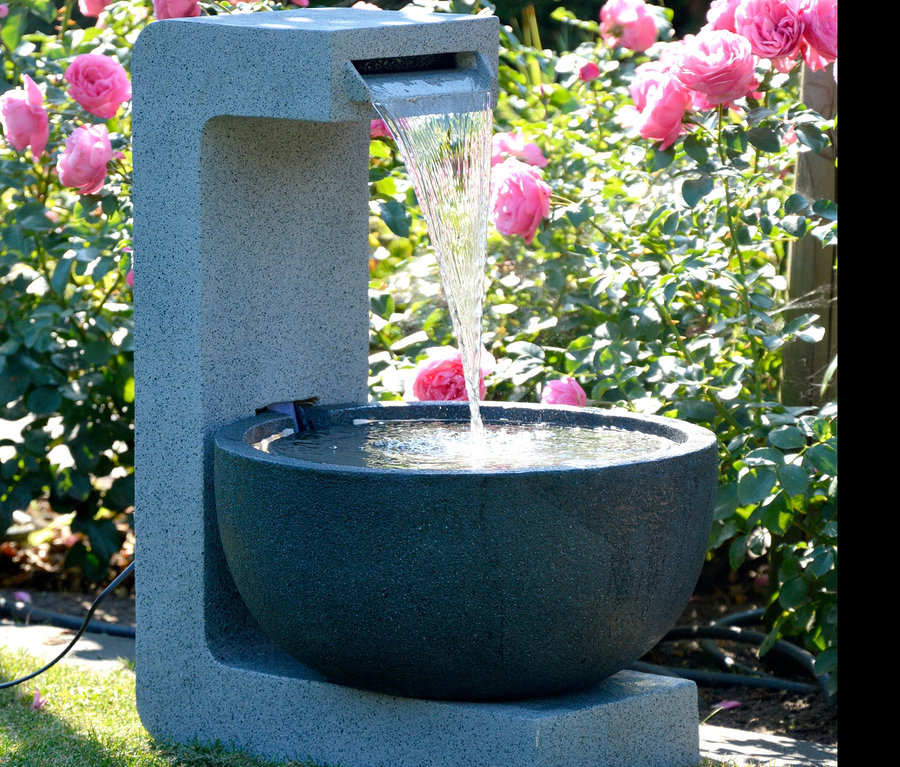 Ein Dobar Gartenbrunnen mit Schale steht auf einem Rasen vor Rosenbüschen.