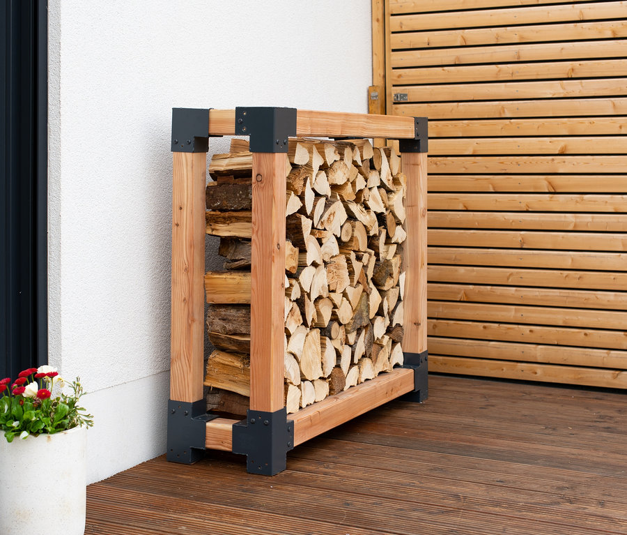 Ein mit Holz gefüllter Kaminholzständer L steht auf einer Holzterrasse neben einer weißen Wand und einer Holztür.