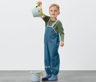 Junge hält einen blauen Eimer über seinen Kopf. Er trägt eine blaue Kleinkind-Regenhose.