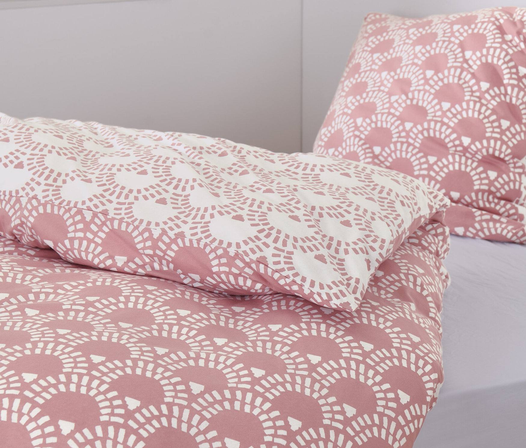 Detailaufnahme von rosafarbener Jersey-Bettwäsche mit Muster. Im Hintergrund sind zwei geflochtene Körbe und Bücher zu sehen.