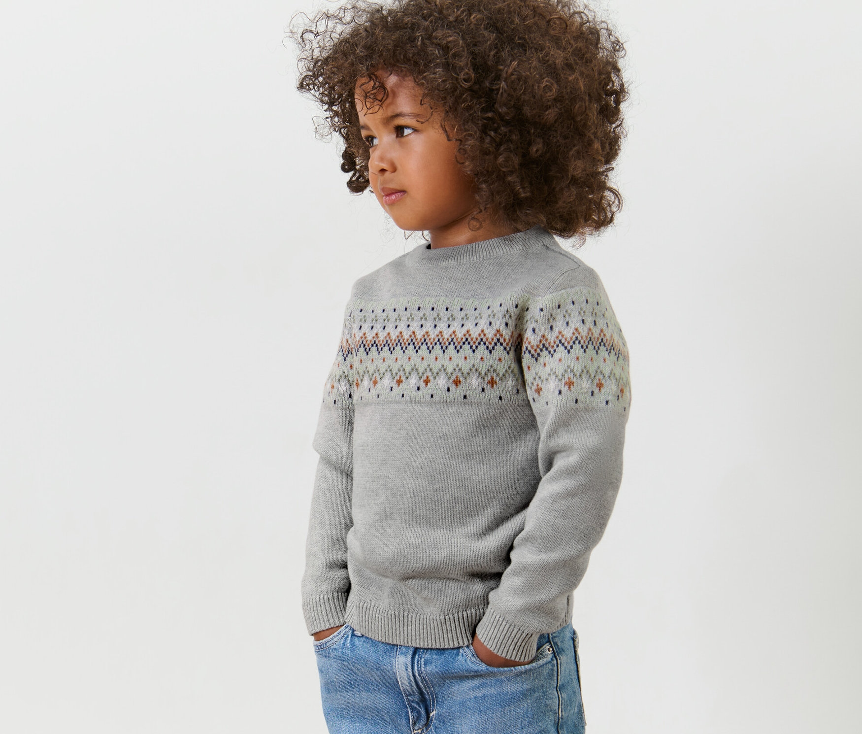 Seitenansicht eines Kindes mit lockigem Haar, das einen grauen Kinder-Strickpullover und Jeans trägt.