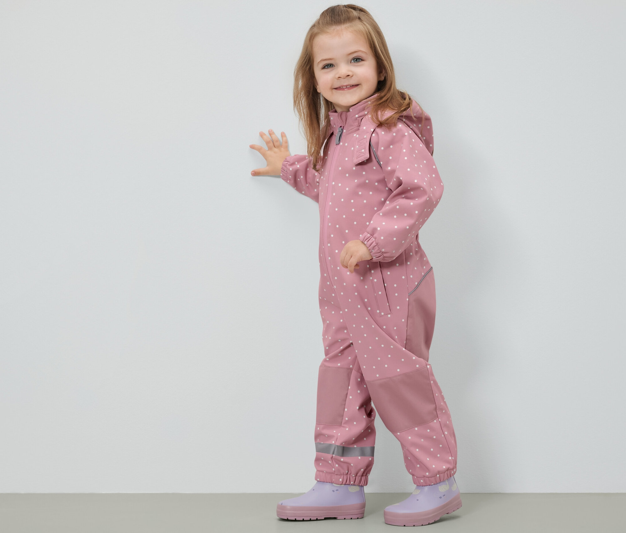 Ein lächelndes Mädchen in einem gepunkteten Kinder-Softshell-Einteiler steht an einer Wand.