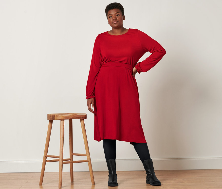 Eine Frau posiert neben einem Holzhocker und trägt ein Jerseykleid in Midi-Länge, Lederboots und eine 2 Paar blickdichte Strumpfhosen, schwarz.