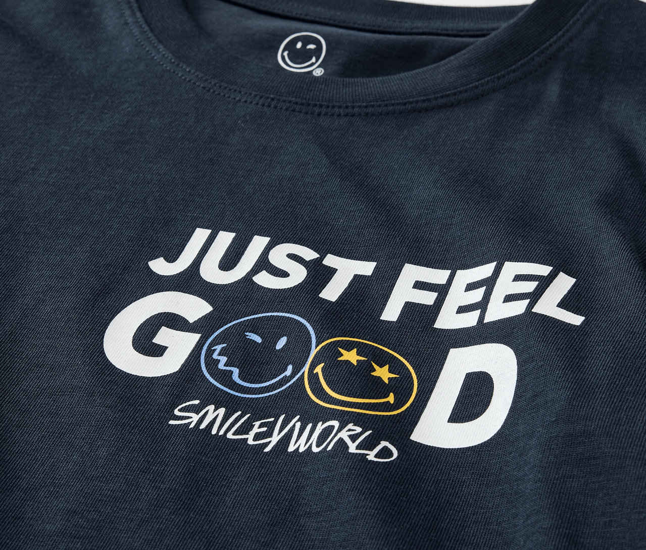 Detailaufnahme eines dunkelblauen T-Shirts mit dem Aufdruck JUST FEEL GOOD und dem SMILEYWORLD-Logo. Kinder-Pyjama SMILEYWORLD®.