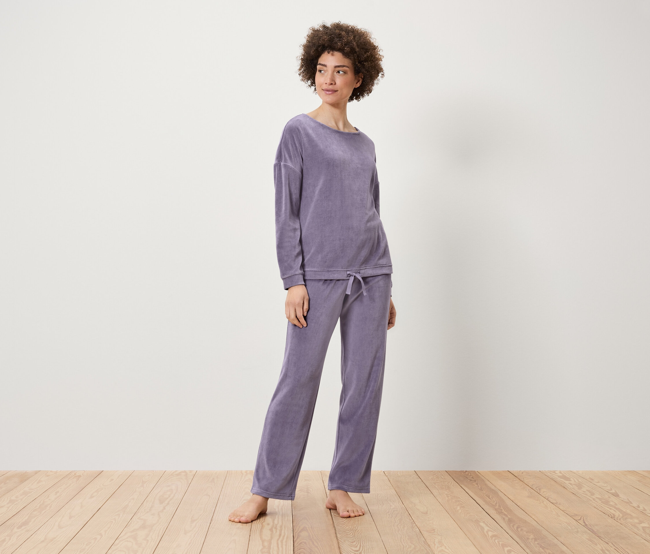 Frau posiert in einem lila Samt-Set aus Sweatshirt und Hose auf einem Holzboden.