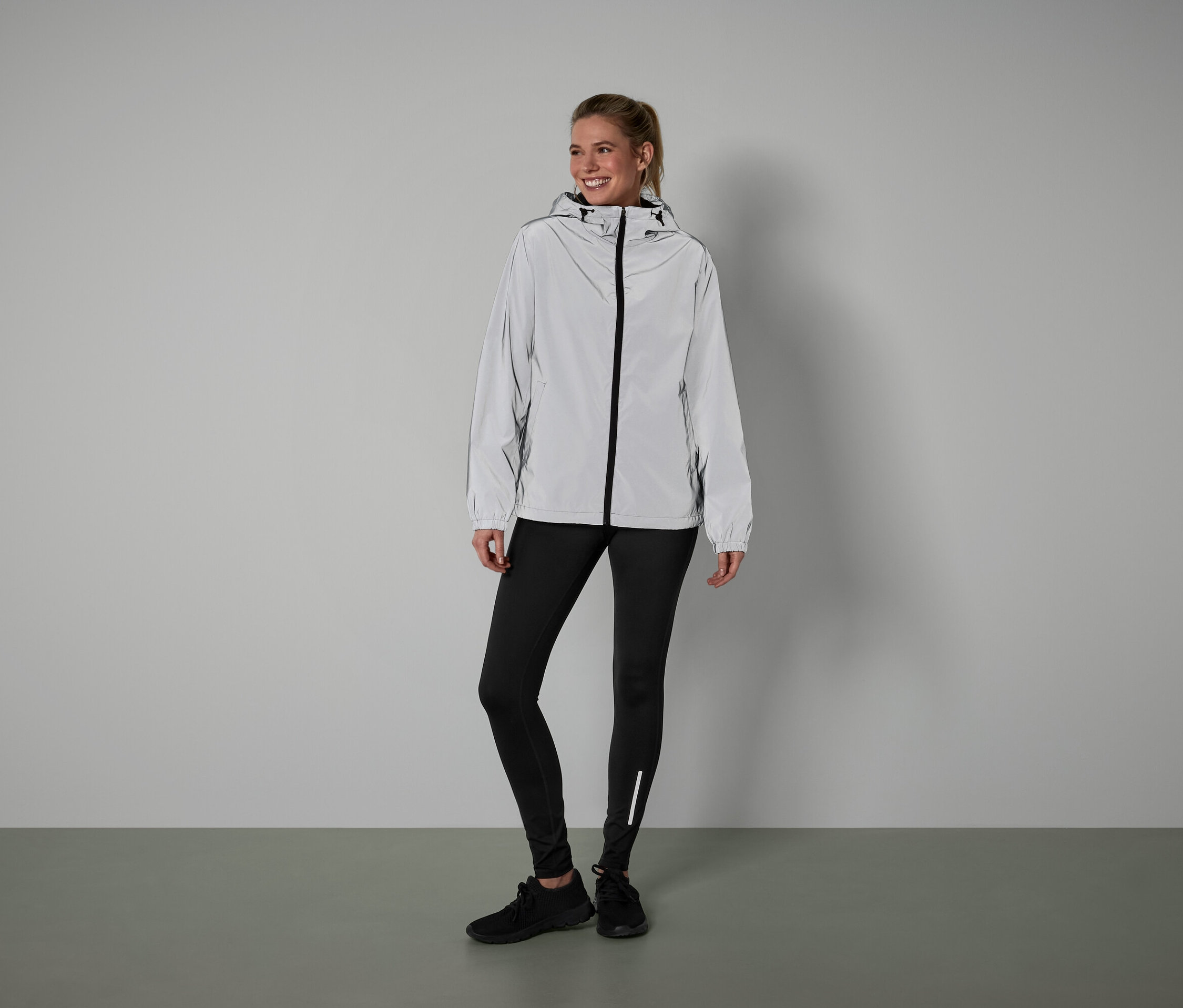 Eine Frau steht und trägt eine silberne Jacke, schwarze Leggings und schwarze Schuhe.
