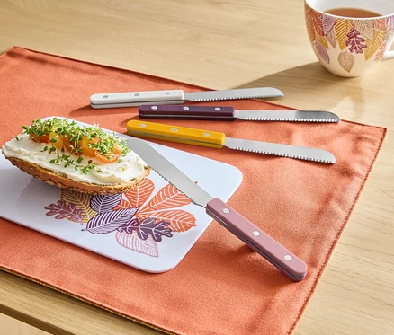 Auf einem orangefarbenen Tischset liegt ein Brot mit Aufstrich auf einem Brettchen mit Blättermuster, daneben liegen vier Messer mit bunten Griffen und im Hintergrund steht eine Tasse Tee.
