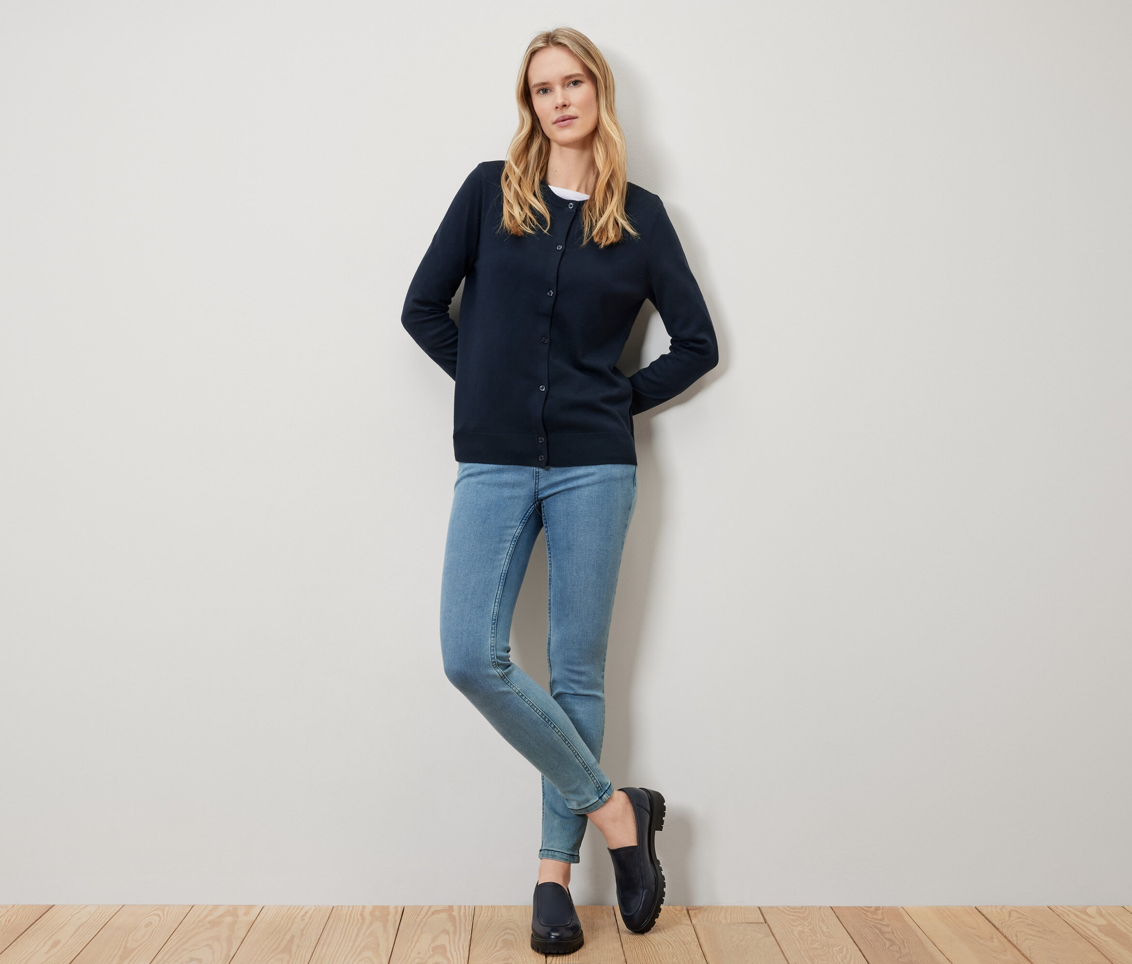 Blonde Frau in blauem Cardigan, Jeans und schwarzen Schuhen steht vor einer weißen Wand.
