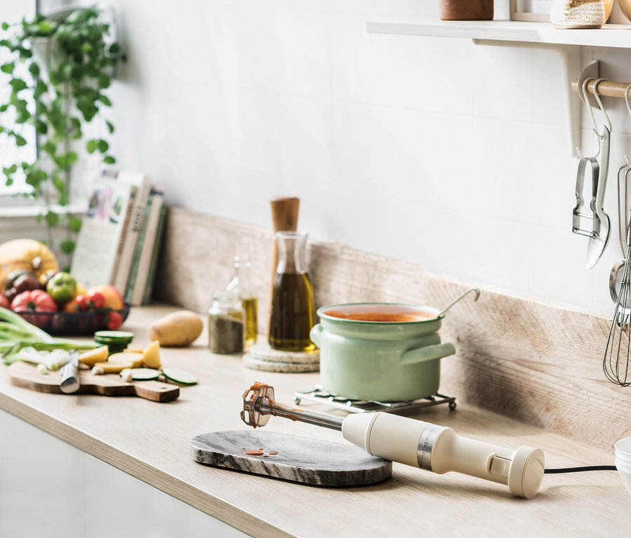 Auf einer Küchentheke stehen ein Mixer auf einem Marmorsockel neben einem grünen Topf mit Suppe, Obst und Gemüse.