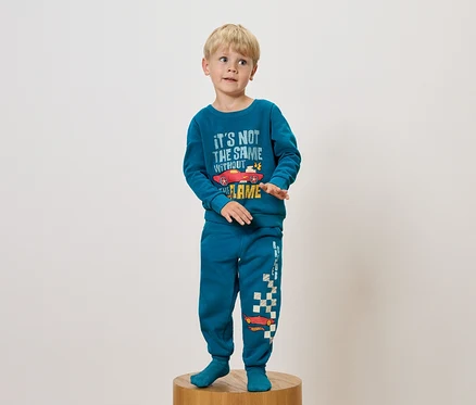 Junge im blauen Pyjama mit Autoaufdruck steht auf einem Holzsockel.