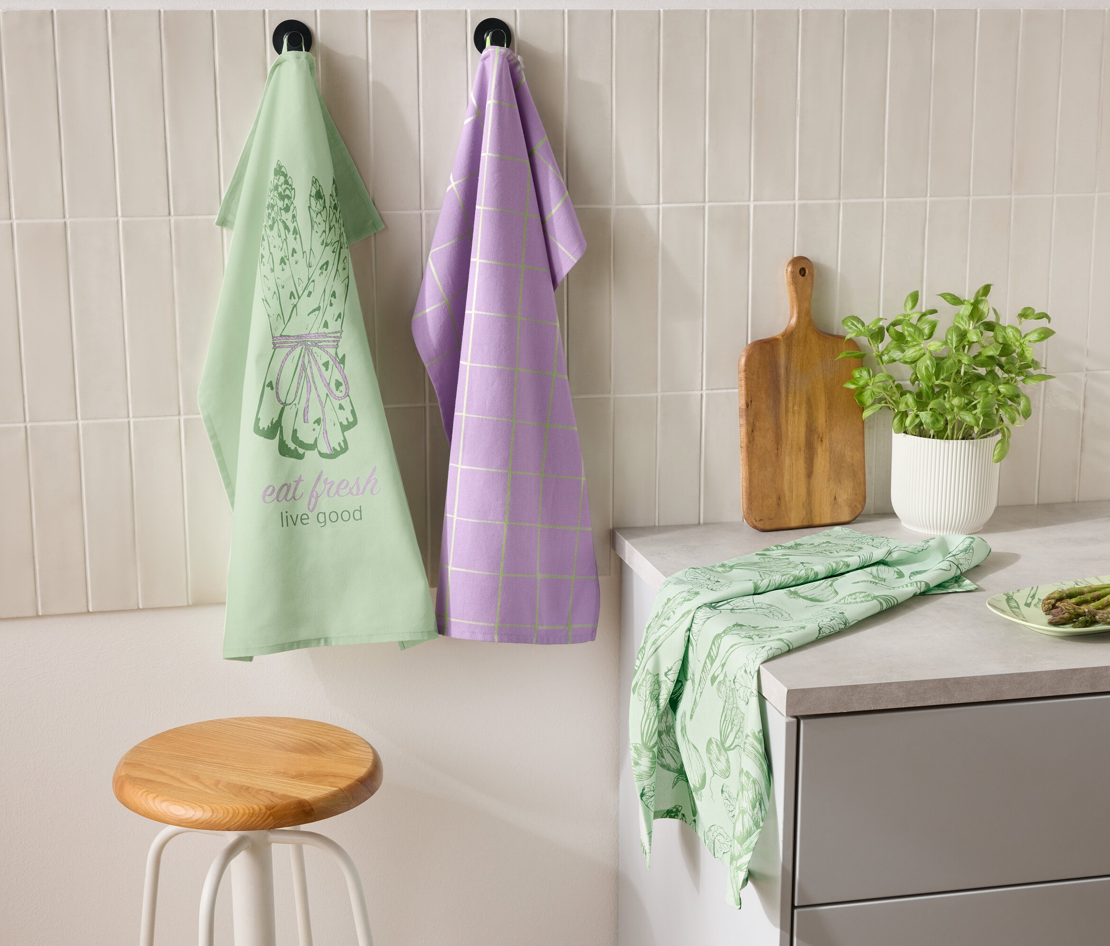 Zwei Geschirrtücher hängen nebeneinander an der Wand, eines grün mit einem Spargelmotiv und dem Schriftzug "eat fresh live good" und das andere lila mit einem Karomuster. Auf der Arbeitsplatte liegen ein grünes Geschirrtuch und ein Teller mit Spargel. Daneben stehen ein Schneidebrett und ein Topf mit Kräutern.