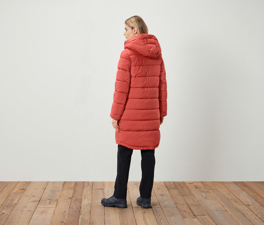 Frau steht mit dem Rücken zum Betrachter und trägt einen roten Steppmantel mit Kapuze, schwarze Hose und dunkle Schuhe.