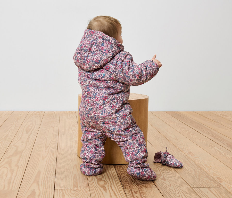 Kleinkind sitzt in einem geblümten Overall auf einem Holzschemel und zeigt mit dem Finger.