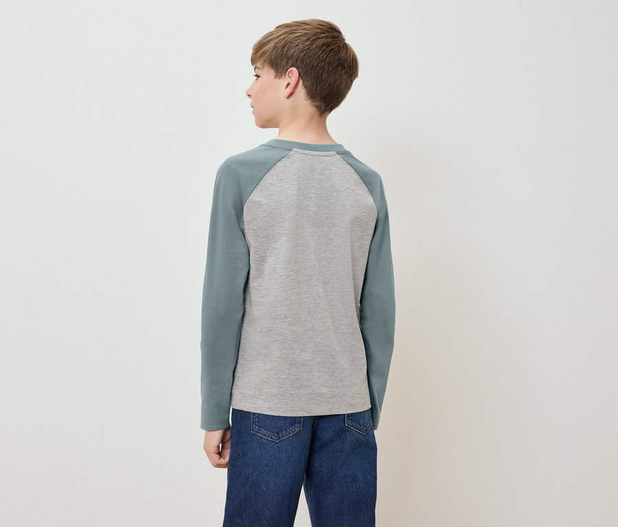 Junge steht mit dem Rücken zur Kamera, trägt ein graues T-Shirt mit langen grünen Ärmeln und blaue Jeans.