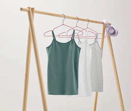 Auf einem hölzernen Kleiderständer hängen drei Tanktops in Grün, Weiß und Grau an rosa Kleiderbügeln. Am Ständer hängt ein Kopfhörer.