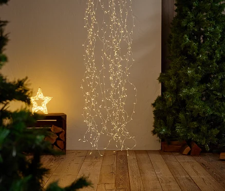 Eine sternförmige Lichterdekoration hängt mit fallenden Lichterketten, daneben steht ein Weihnachtsbaum und ein leuchtender Stern.