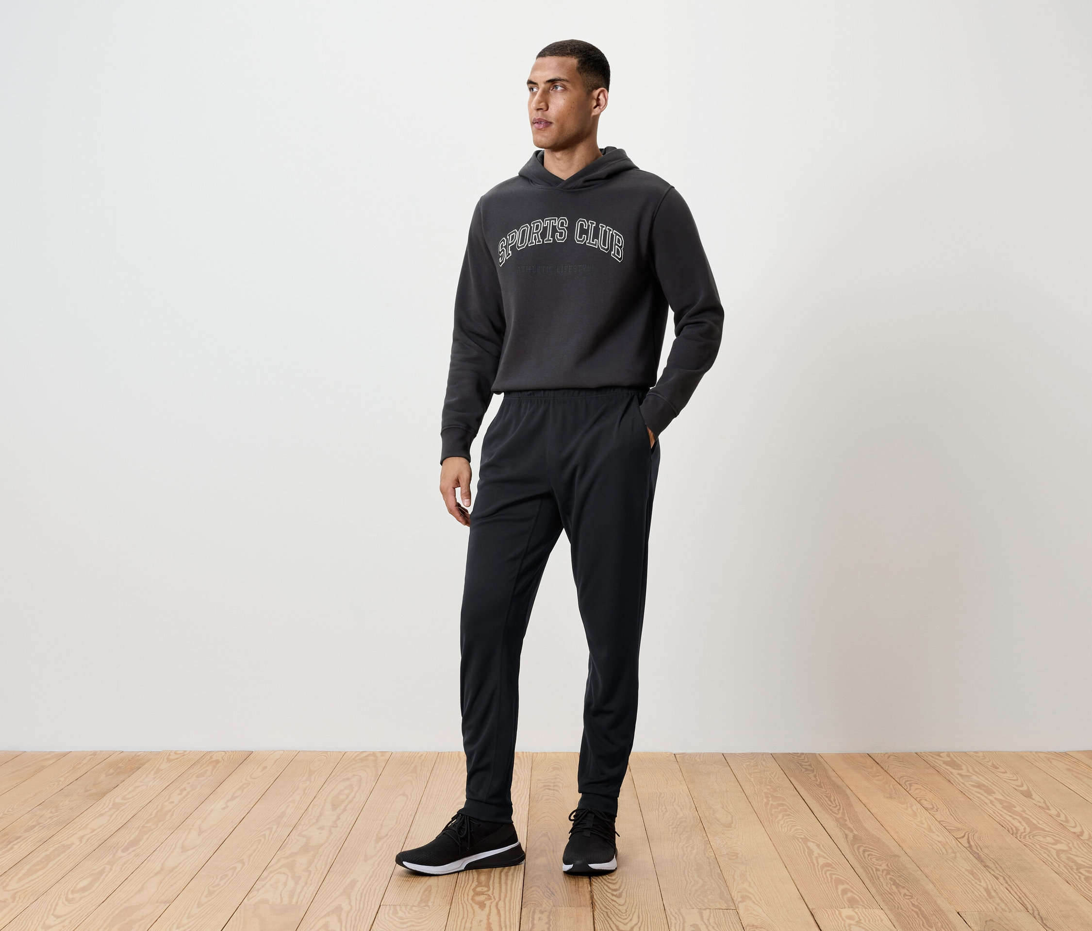 Mann steht auf einem Holzboden und trägt eine schwarze Trainingshose, einen Kapuzenpullover und Turnschuhe.