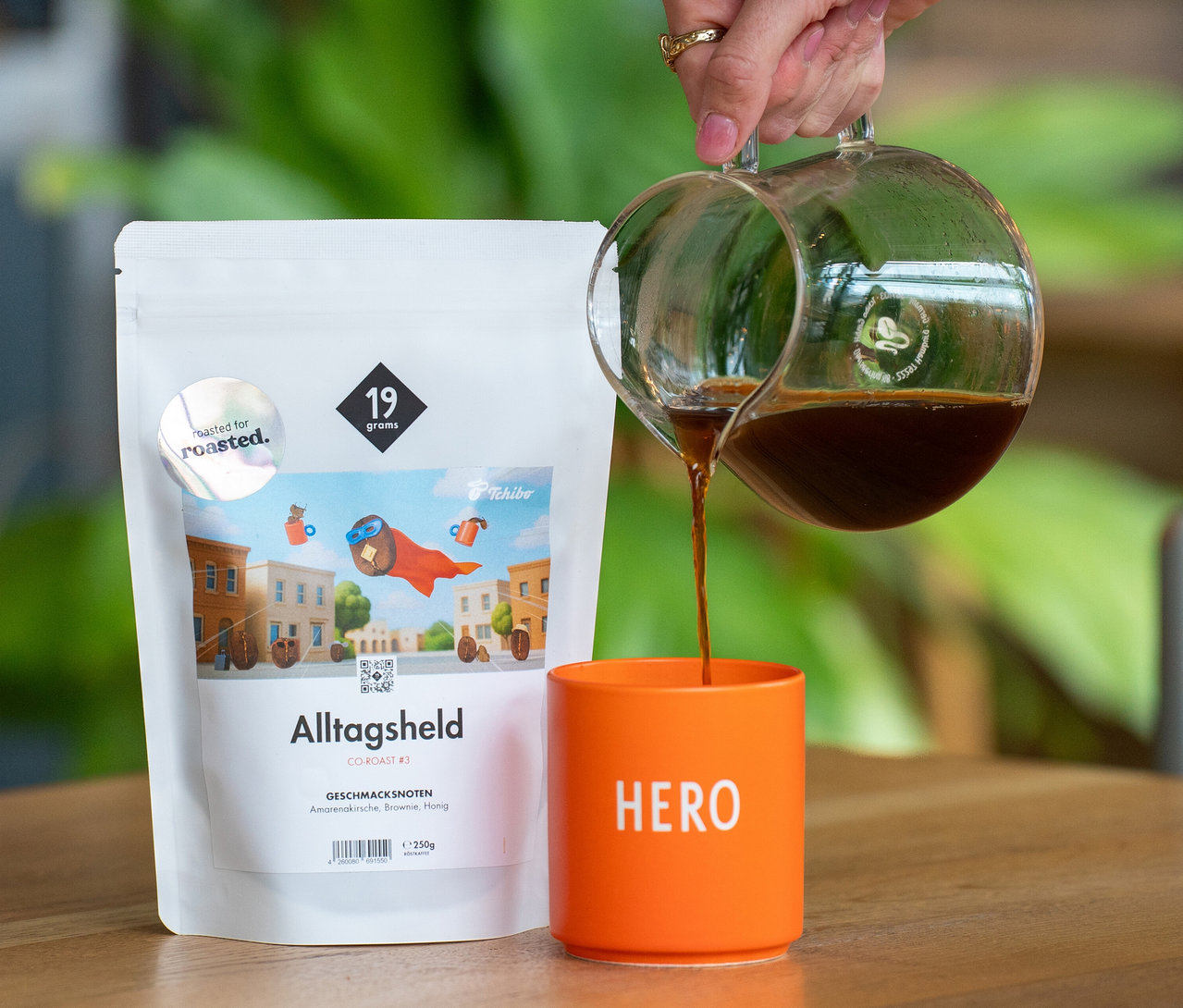 Eine Hand gießt Kaffee aus einer Glaskanne in eine orangefarbene Tasse mit der Aufschrift „HERO“. Daneben steht eine weiße Packung 19grams x roasted. Alltagsheld Co-Roast #3 Omni-Roast mit 250 g ganzen Bohnen auf einem Holztisch.