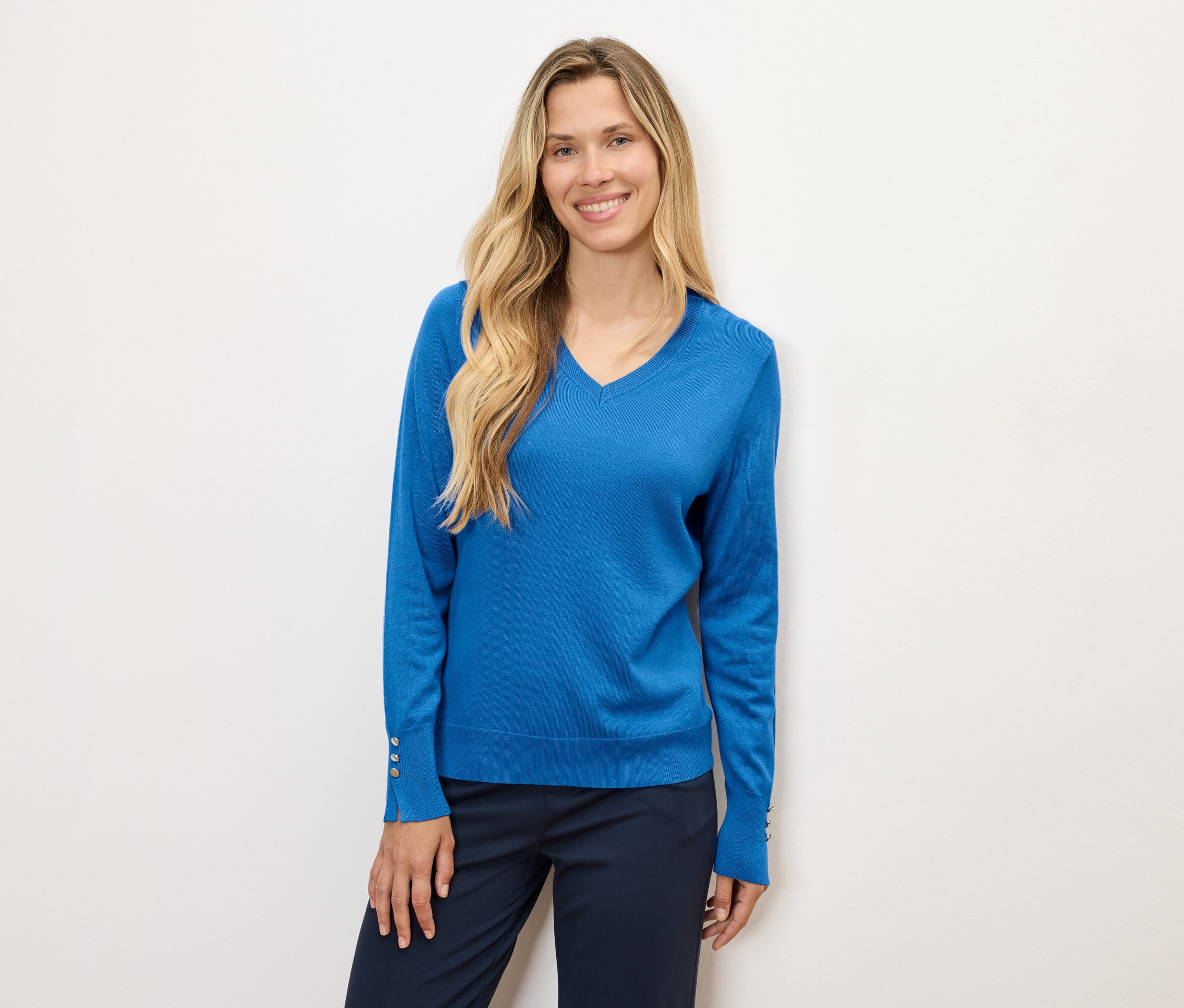 Eine Frau mit langen blonden Haaren posiert in einem blauen Pullover und dunkelblauen Hosen.