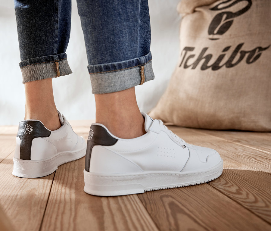 Beine einer Frau in Jeans und weißen Tchibo x VLACE Coffee Lover Sneakern stehen auf einem Holzboden neben einem Jutesack mit Tchibo-Logo.
