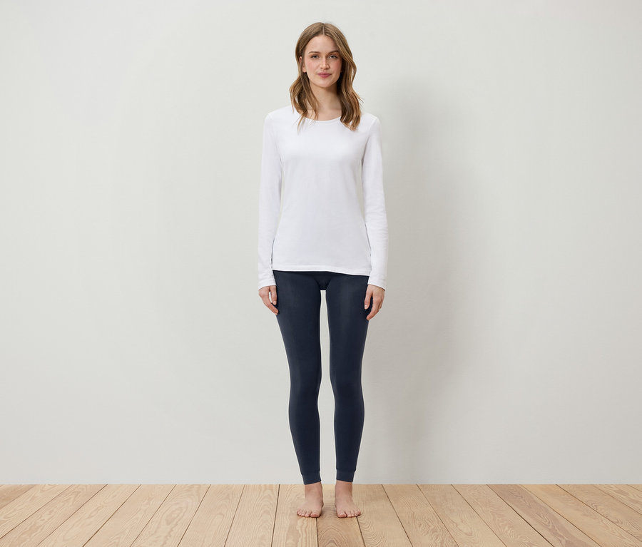 Eine Frau steht auf einem Holzboden und trägt ein weißes Langarmshirt und dunkelblaue Leggings.