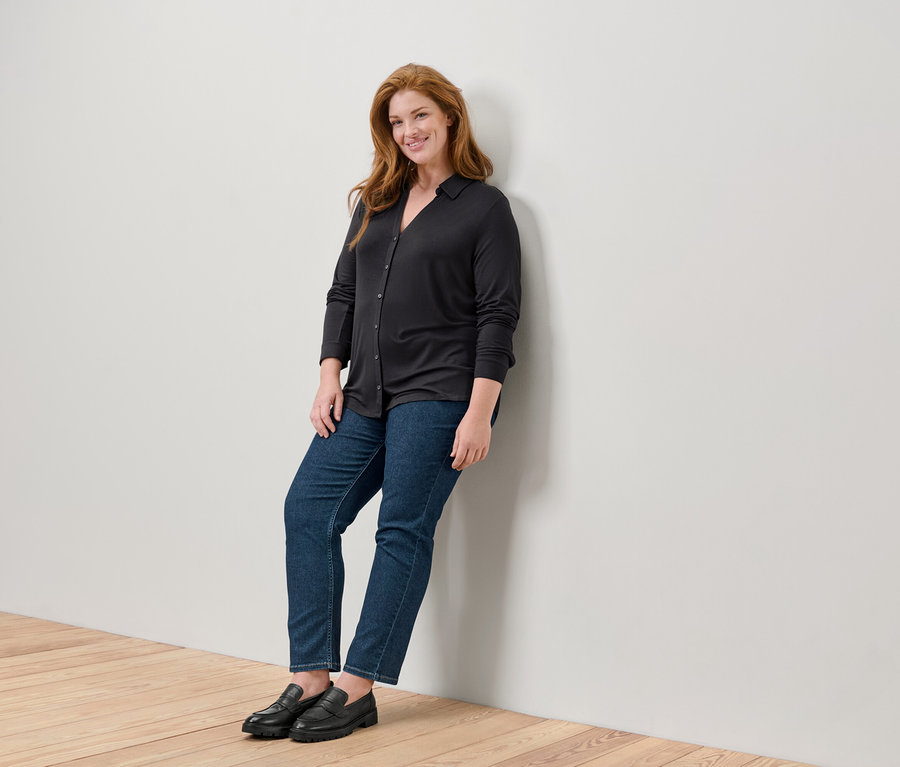 Eine Frau mit langen roten Haaren steht an eine Wand gelehnt und trägt einen schwarzen Pullover, Jeans und schwarze Slipper.