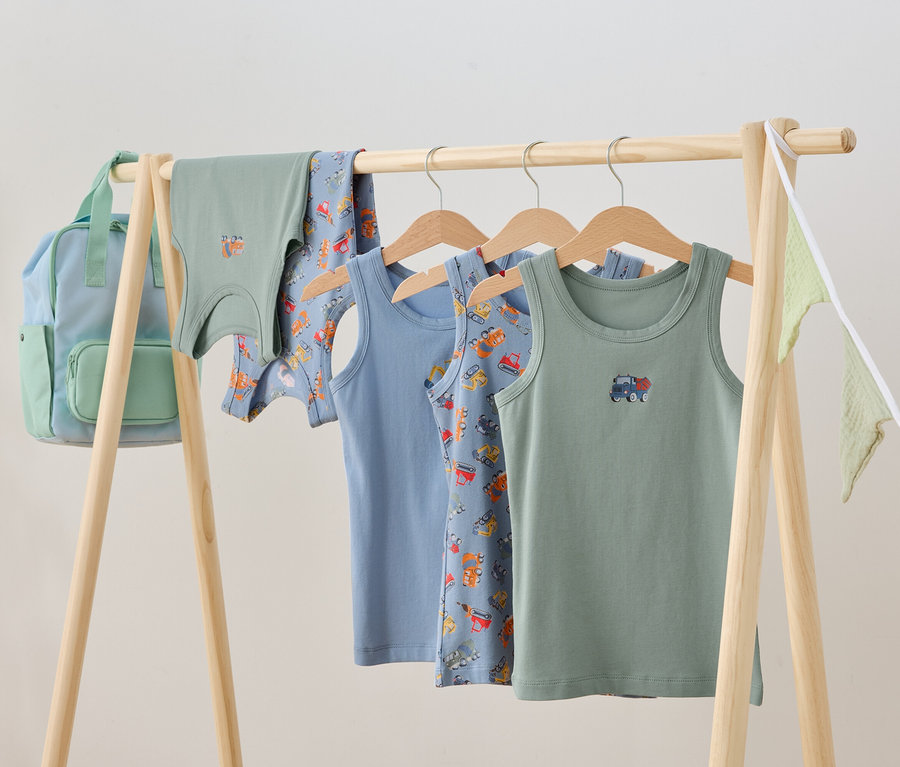 Kinderkleidung hängt auf einem Holzständer, darunter Unterhemden und ein Rucksack.