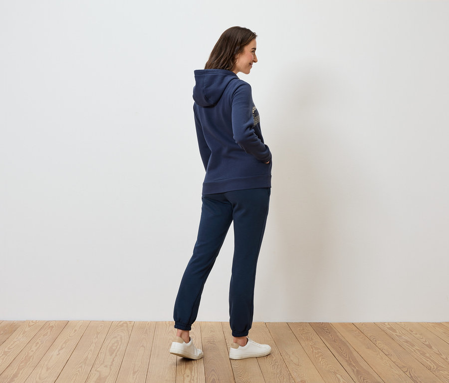 Eine Frau in marineblauem Hoodie und Trainingshose steht seitlich auf einem Holzboden.