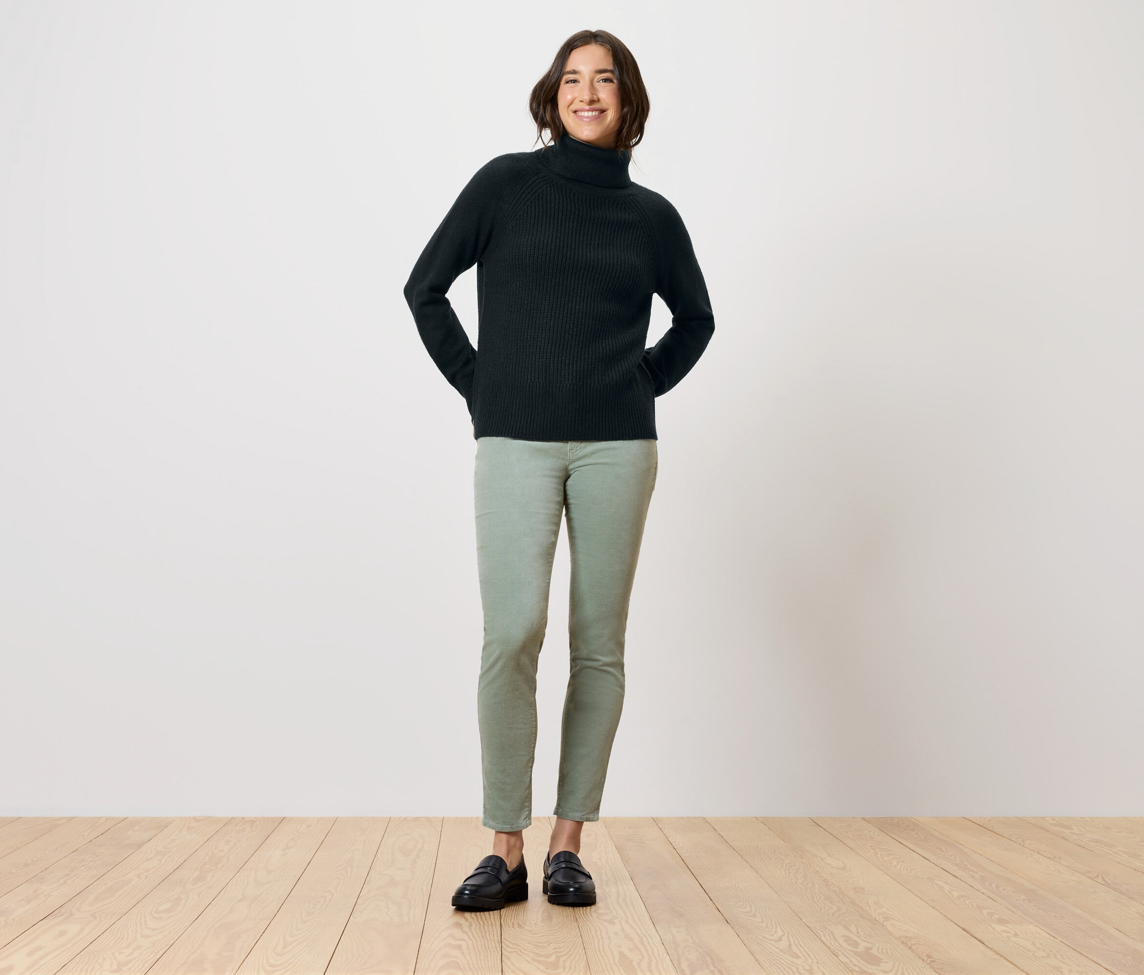 Eine Frau steht mit den Händen hinter dem Rücken, sie trägt einen schwarzen Rollkragenpullover, grüne Hosen und schwarze Schuhe.